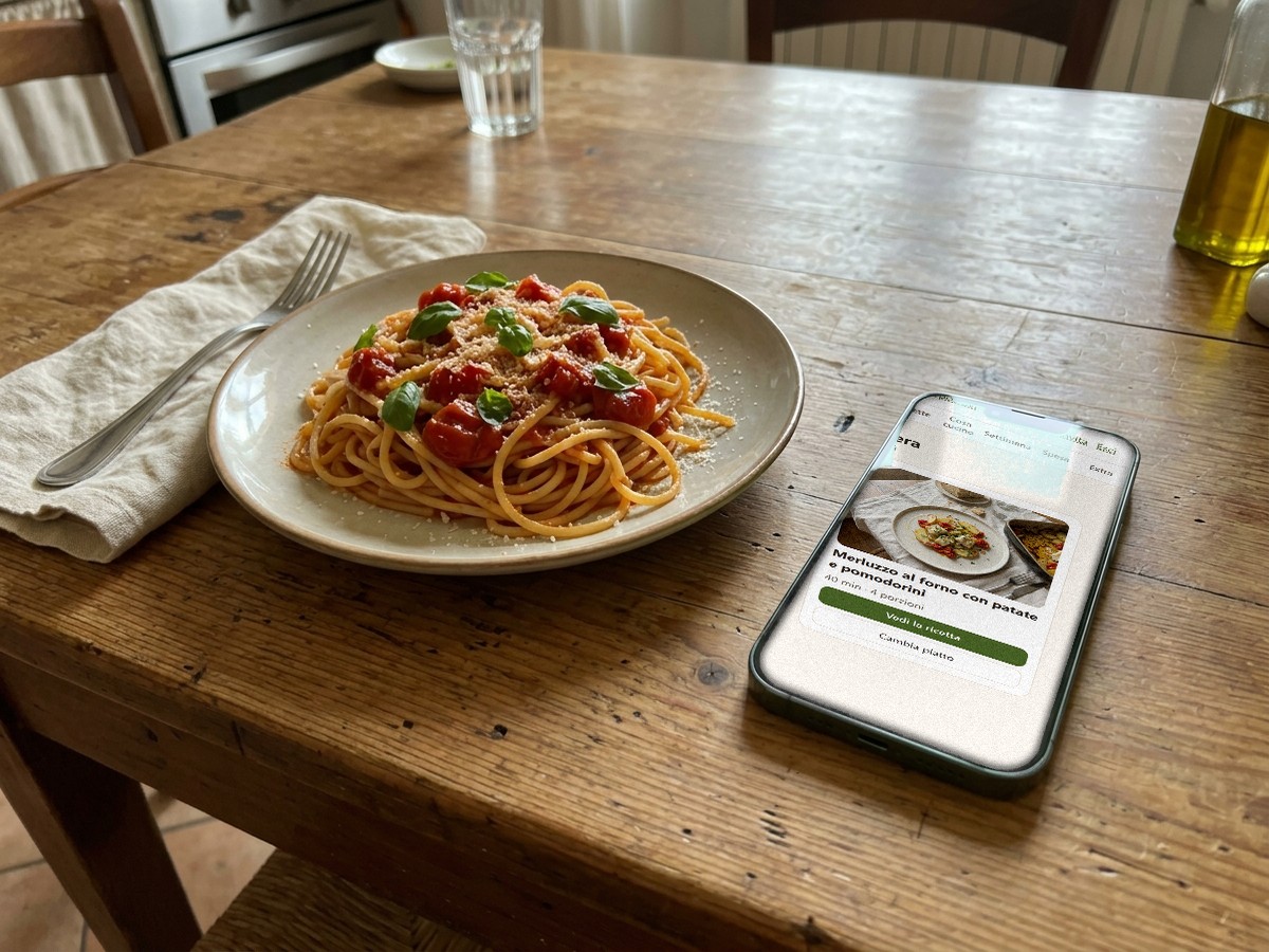 Il telefono con la schermata «Stasera» dell'app, accanto a un piatto di pasta al pomodoro fresco e basilico